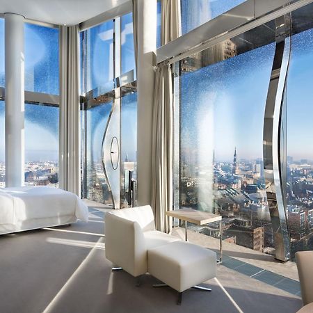The Westin Hamburg Elbphilharmonie Hotel Ngoại thất bức ảnh The photo shows a modern hotel room with a stunning, panoramic view through large, uniquely designed windows. The room features a sleek, minimalist aesthetic with light-colored furnishings, including a white bed and a couple of chairs. The walls are 
