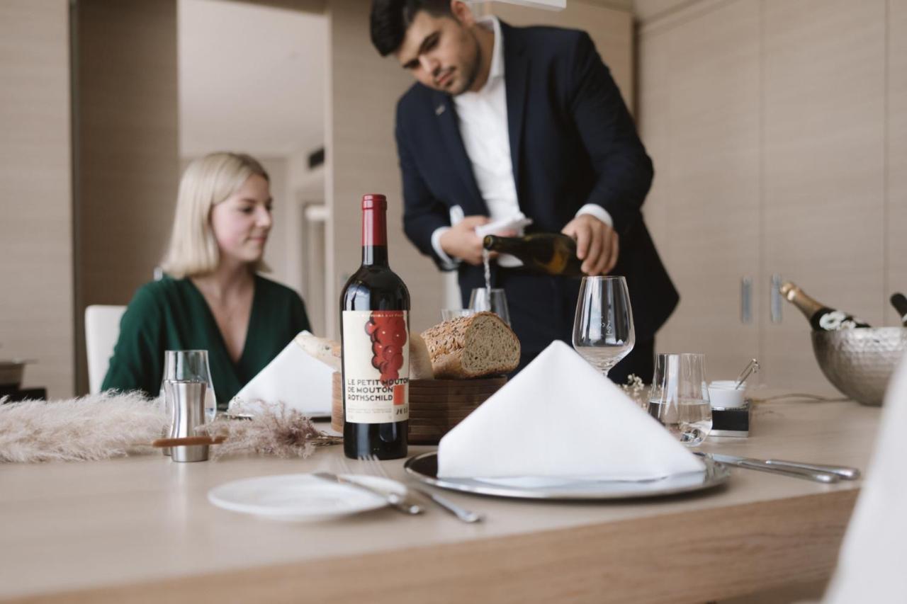 The Westin Hamburg Elbphilharmonie Hotel Ngoại thất bức ảnh The photo shows a dining scene featuring a well-dressed man pouring wine into a glass while a woman sits at the table. The table is elegantly set with a folded napkin, wine glasses, and a bottle of red wine placed prominently. There is also a loaf of
