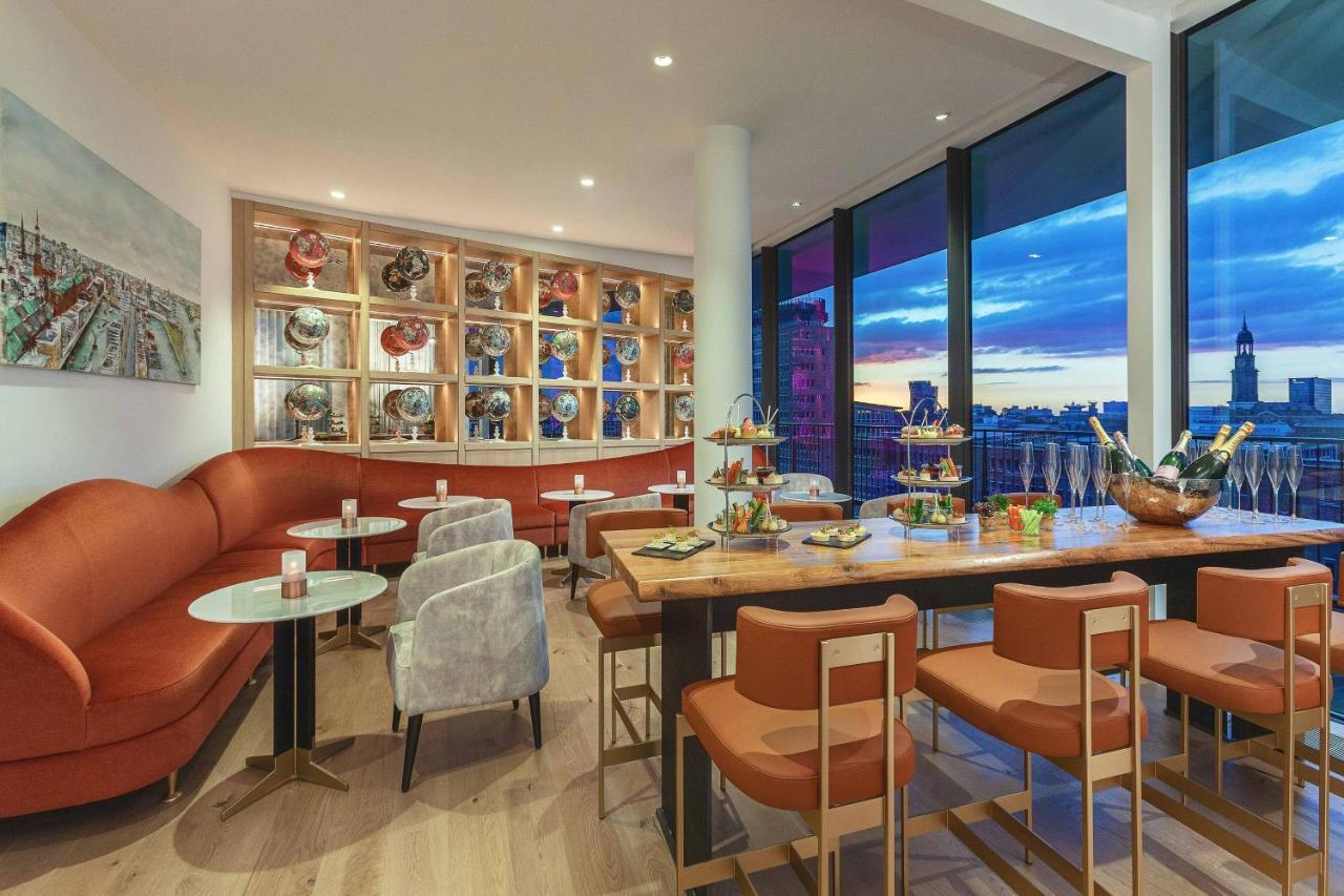 The Westin Hamburg Elbphilharmonie Hotel Ngoại thất bức ảnh The photo shows a modern and stylish restaurant or café interior. It features a combination of comfortable seating, including a curved, orange sofa and gray chairs. The tables are round with light-colored tops, and there are several tiered stands dis