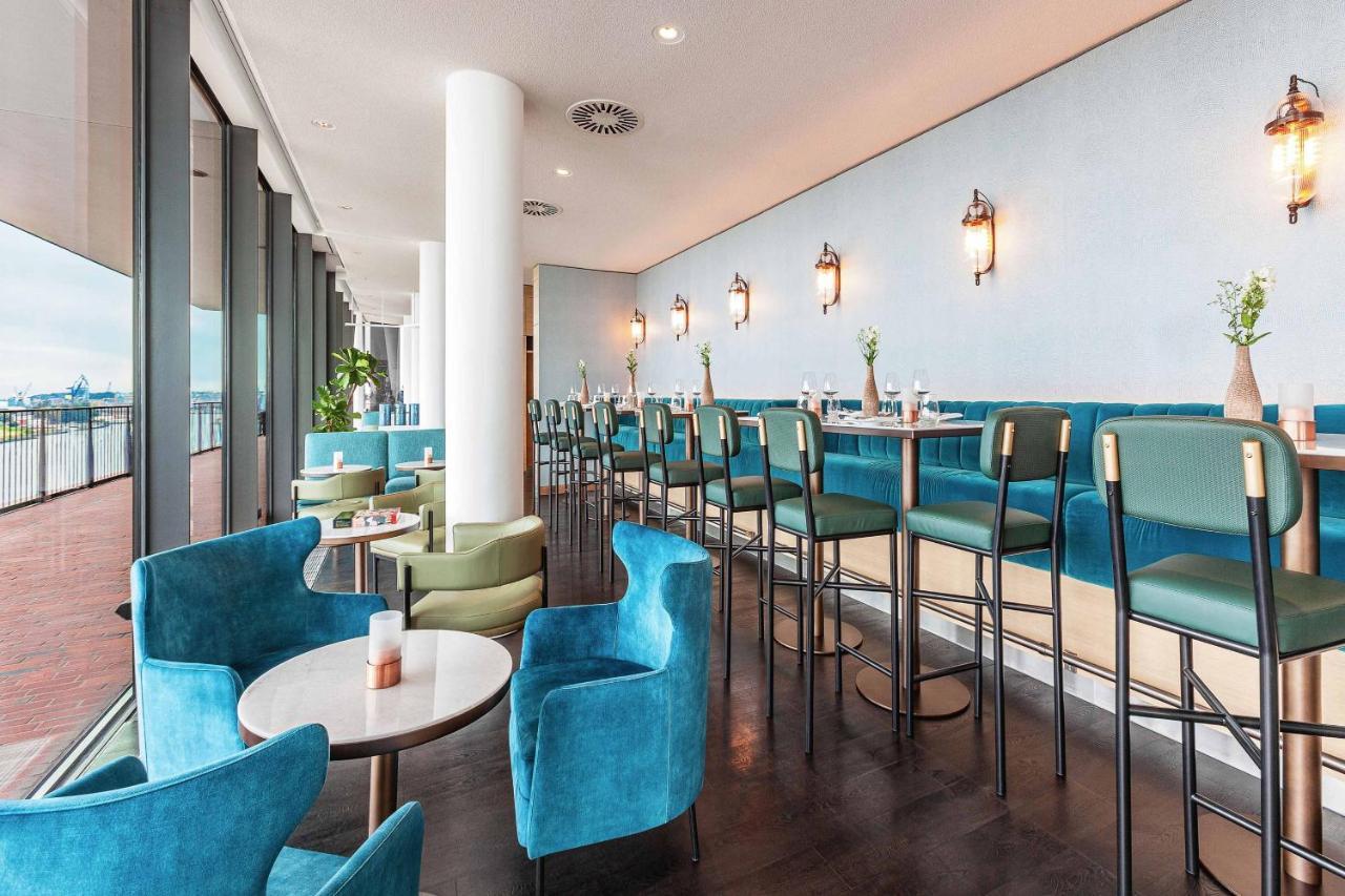 The Westin Hamburg Elbphilharmonie Hotel Ngoại thất bức ảnh The photo shows a modern and stylish restaurant or café interior. It features a long row of high-backed bar stools at a counter, complemented by plush seating in the form of rounded chairs and small tables arranged throughout the space. The color sch