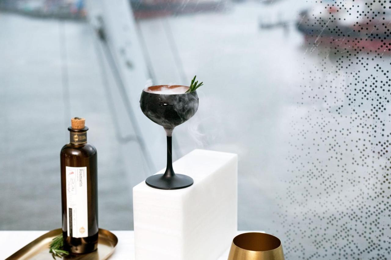 The Westin Hamburg Elbphilharmonie Hotel Ngoại thất bức ảnh The photo features a stylish cocktail elegantly presented on a white pedestal. The cocktail is served in a sophisticated black stem glass, topped with a garnish of rosemary. Beside it, there is a dark glass bottle, likely containing a spirit or mixer