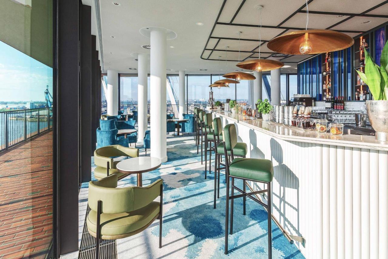 The Westin Hamburg Elbphilharmonie Hotel Ngoại thất bức ảnh The photo depicts a modern bar or lounge area with a stylish design. There are high-backed green chairs surrounding small circular tables, arranged in an open space that has large windows showcasing a view of the outside. The bar area is sleek, featu