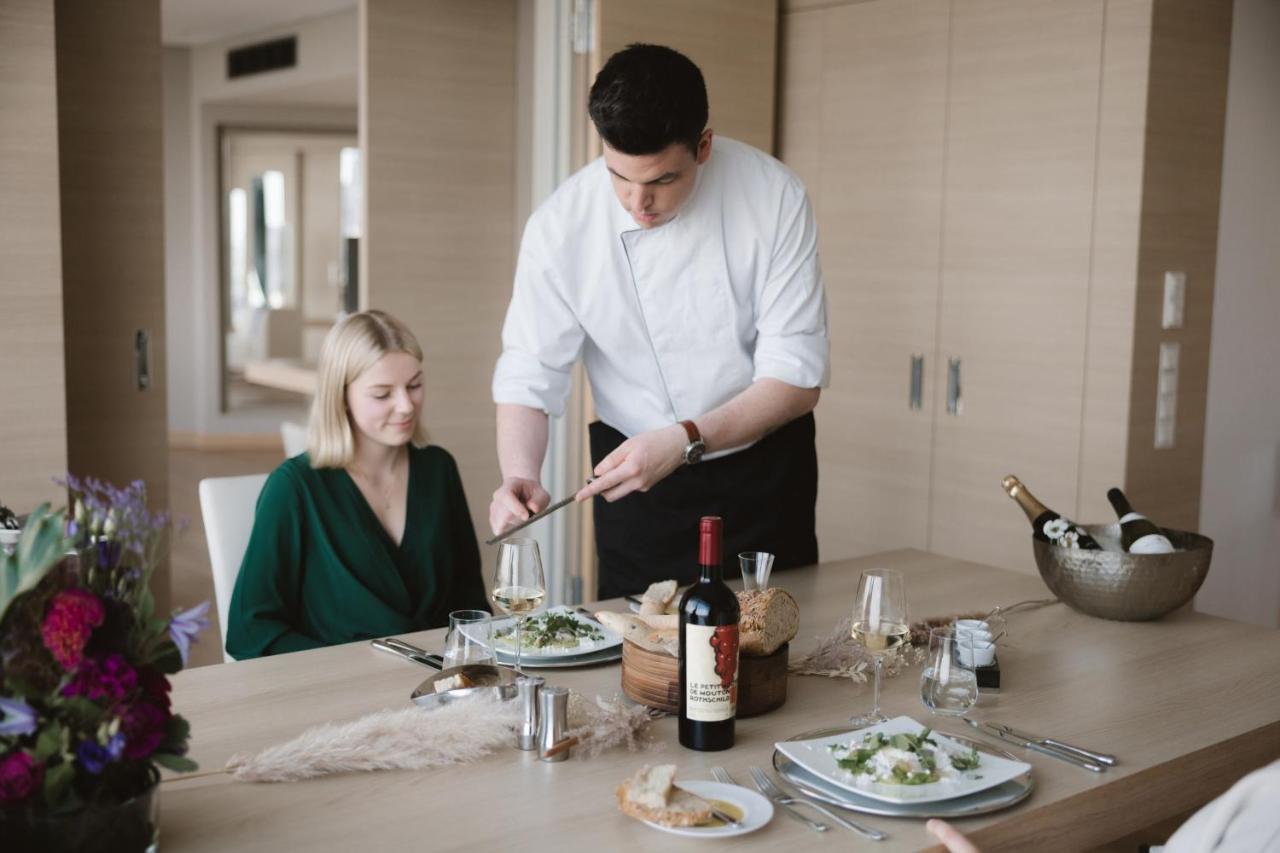 The Westin Hamburg Elbphilharmonie Hotel Ngoại thất bức ảnh The photo depicts a dining scene in a modern setting. A man, dressed in a chef's attire, is serving food to a woman seated at the table. The woman is wearing a green outfit and appears to be enjoying the meal. The table is elegantly set with plates o