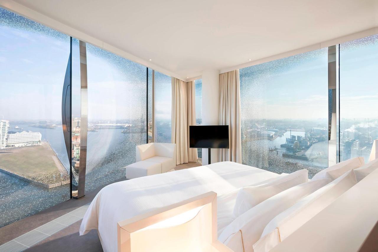 The Westin Hamburg Elbphilharmonie Hotel Ngoại thất bức ảnh The photo shows a modern hotel room featuring a large bed with white linens and a minimalist design. It has expansive floor-to-ceiling windows that offer a panoramic view of the landscape outside, likely a city or waterfront setting. The room is deco