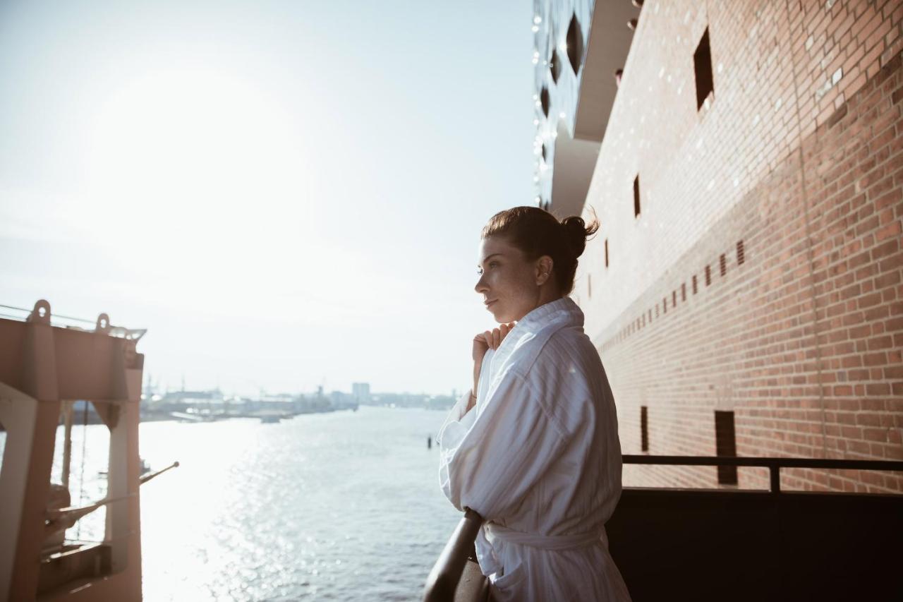 The Westin Hamburg Elbphilharmonie Hotel Ngoại thất bức ảnh The photo depicts a person standing on a balcony, looking out over a body of water. The individual is wearing a white bathrobe, and their hair is styled in a bun. The background features a scenic view with sunlight illuminating the scene, creating a 