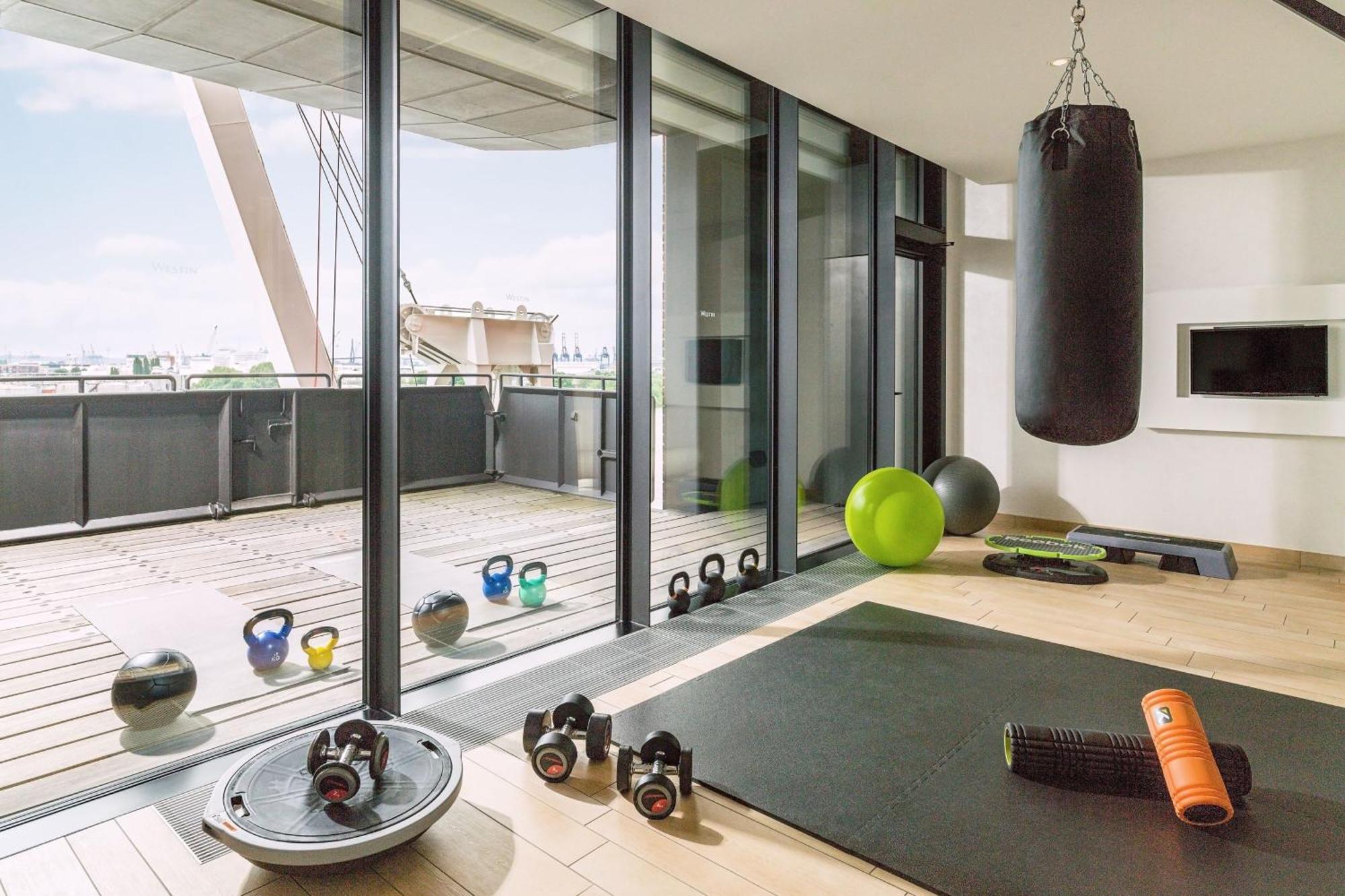 The Westin Hamburg Elbphilharmonie Hotel Ngoại thất bức ảnh The photo shows a modern home gym with large glass windows that provide a view to the outside. Inside, there are various fitness equipment items, including a heavy punching bag hanging from the ceiling, kettlebells, a stability ball, an exercise mat,