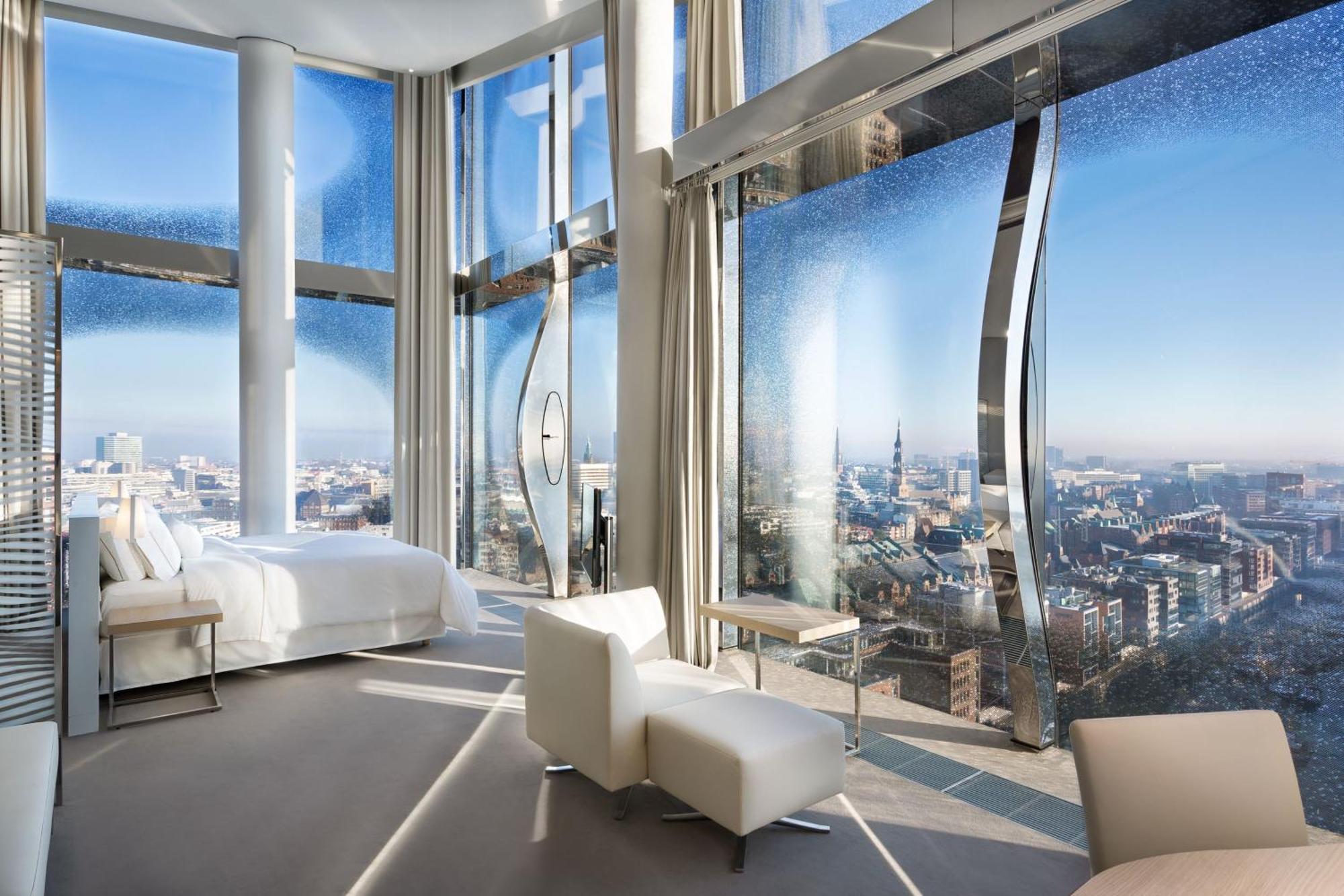 The Westin Hamburg Elbphilharmonie Hotel Ngoại thất bức ảnh The photo shows a modern hotel room with a stunning, panoramic view through large, uniquely designed windows. The room features a sleek, minimalist aesthetic with light-colored furnishings, including a white bed and a couple of chairs. The walls are 